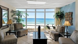 A spacious living room with a large window overlooking a beach, furnished with sofas and a coffee table. - 2 Bedroom in Ocean Villa Resort -  Beautifully decorated and appointed condo with amazing views! (Panama City Beach)