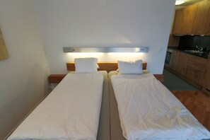 Une chambre double avec deux lits simples, une tête de lit en bois, une petite table de chevet et une kitchenette dotée de meubles en bois et d’un plan de travail noir.