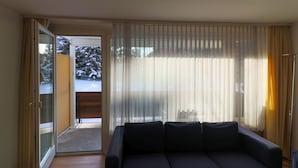 Una sala de estar con un sofá oscuro, un ventanal corredero que da a un balcón y cortinas de tela ligera.