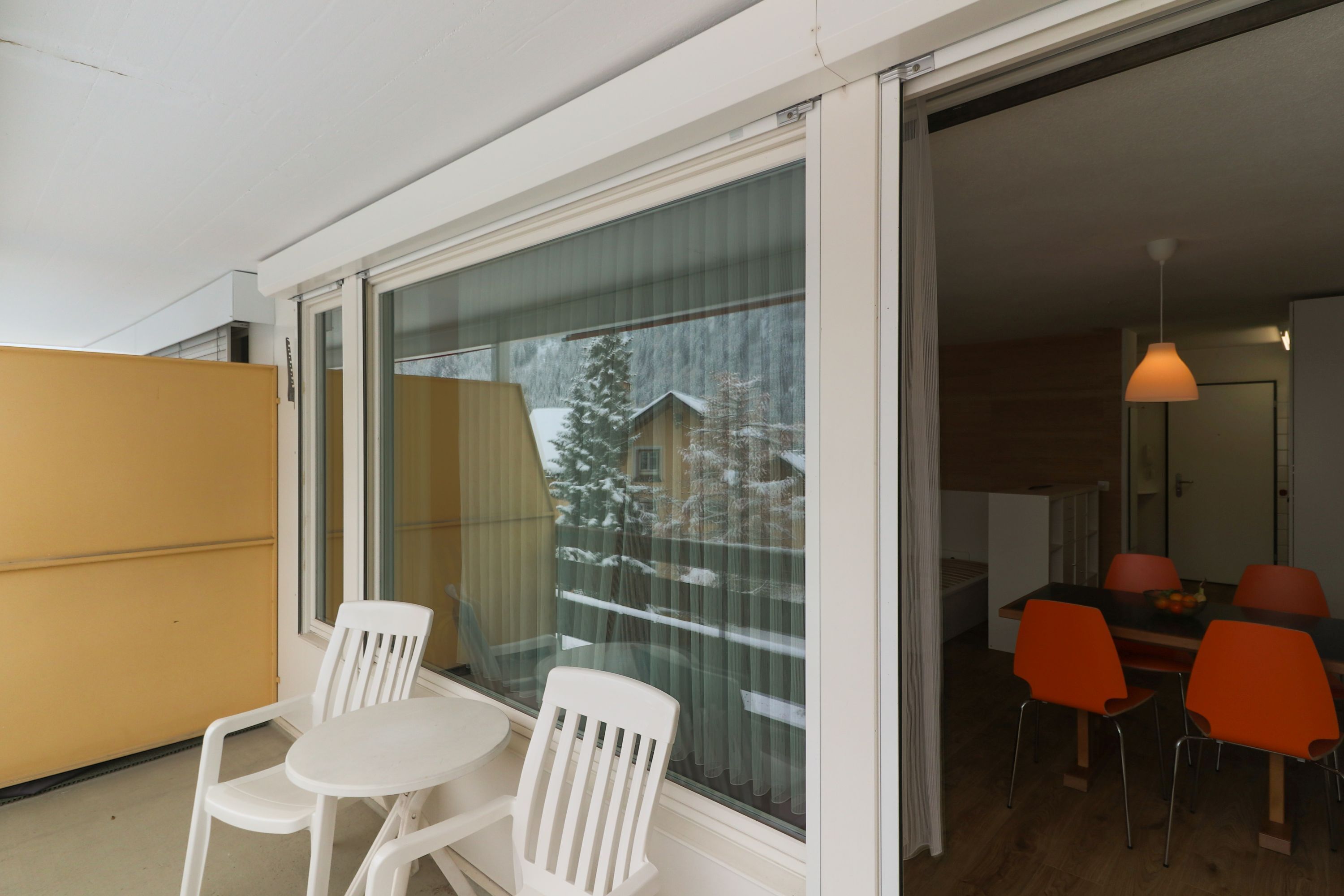 A balcony with white chairs, a wooden table, and a view of a snowy landscape through a glass door.