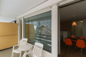 Un balcon avec des chaises blanches, une table en bois et une vue sur un paysage enneigé, visible à travers une porte vitrée.