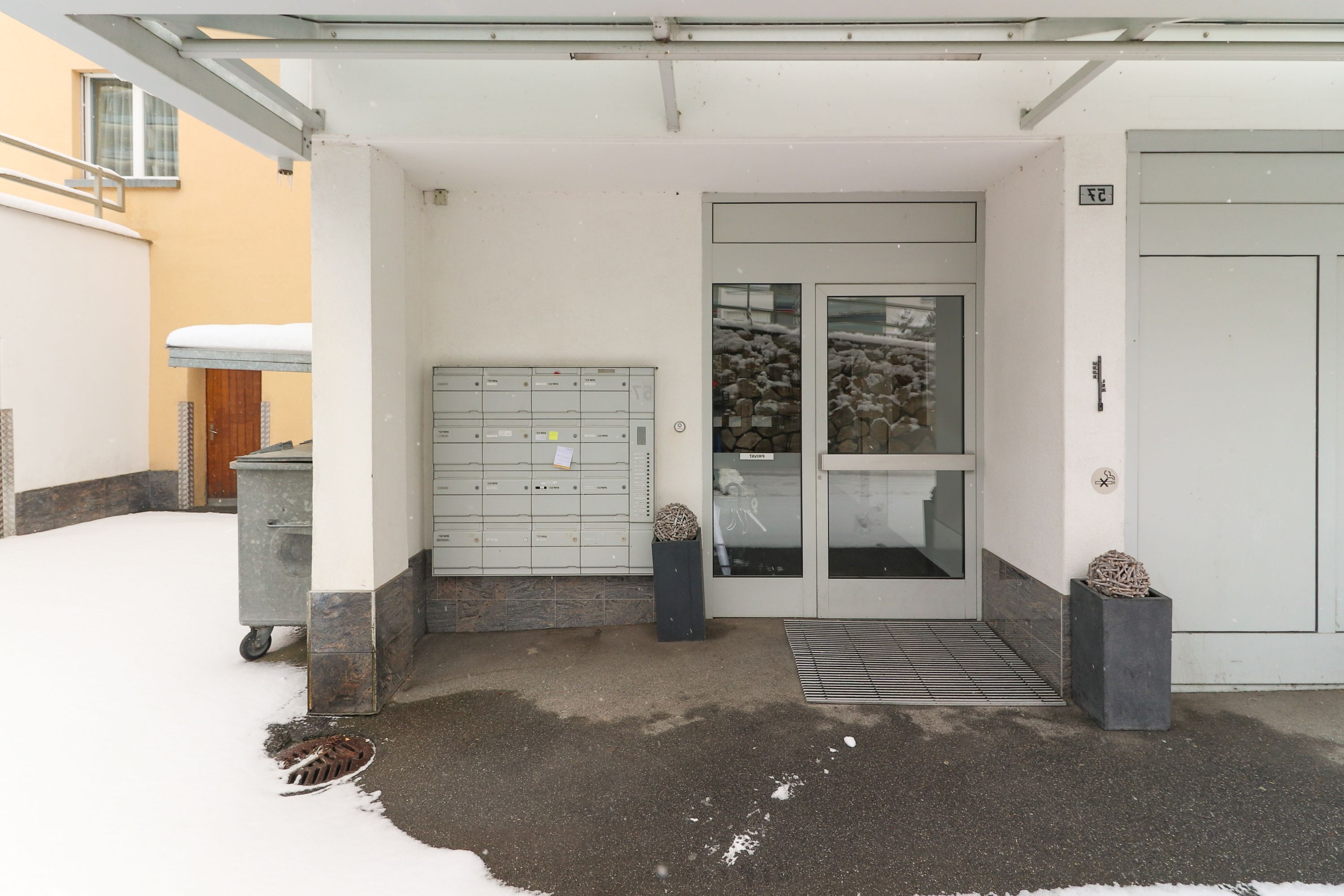 A building entrance with a snow-covered ground, a metal door, and a small step leading up to the entrance.