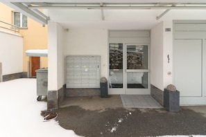 Une entrée d’immeuble avec un sol recouvert de neige, une porte en métal et une petite marche menant à l’entrée.