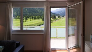 A room with a large window offering a view of a green landscape with trees and mountains. - Apartments Schmid (Davos)