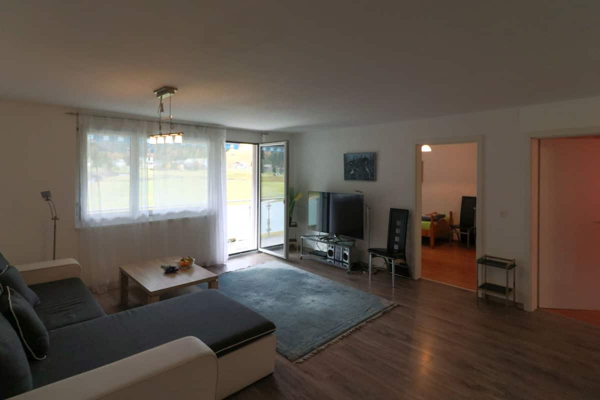 A living room with a sofa, a television, and a small table. There is a sliding glass door leading to a balcony.