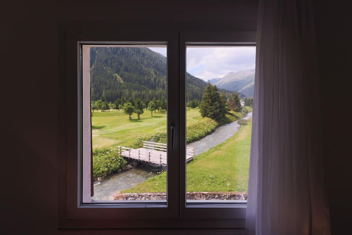A room with a large window offering a view of a green landscape with a river, trees, and mountains.