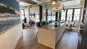 A modern kitchen with a central island, wooden flooring, and large windows providing natural light.