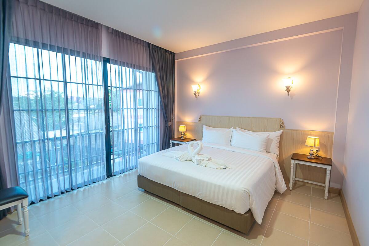 A hotel room with a large bed, two bedside tables with lamps, a bench, and a view through a large window with curtains.