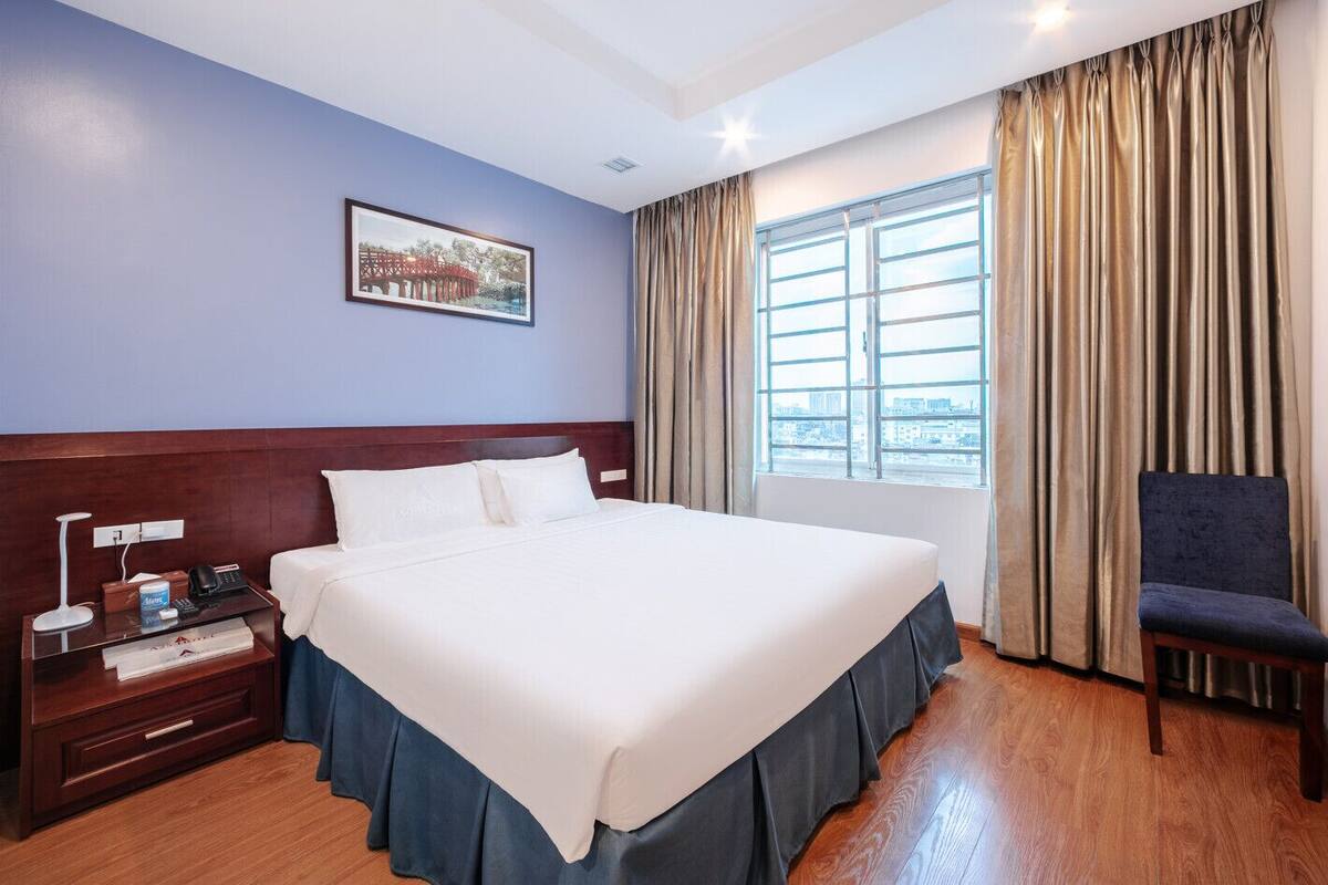 Une chambre d’hôtel avec un grand lit, une table de chevet avec une lampe, une chaise, une fenêtre avec des rideaux et un tableau encadré au mur.