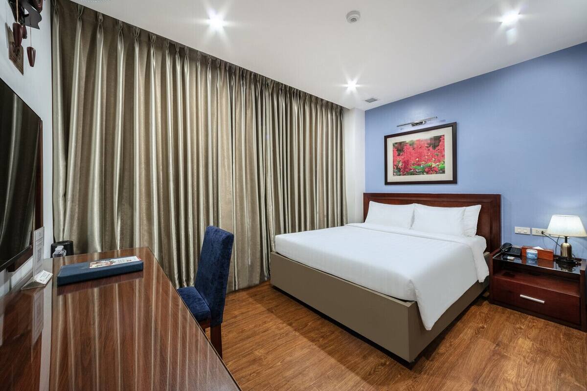 Une chambre d’hôtel avec un lit, un bureau, une chaise, une table de chevet et un tableau au mur.