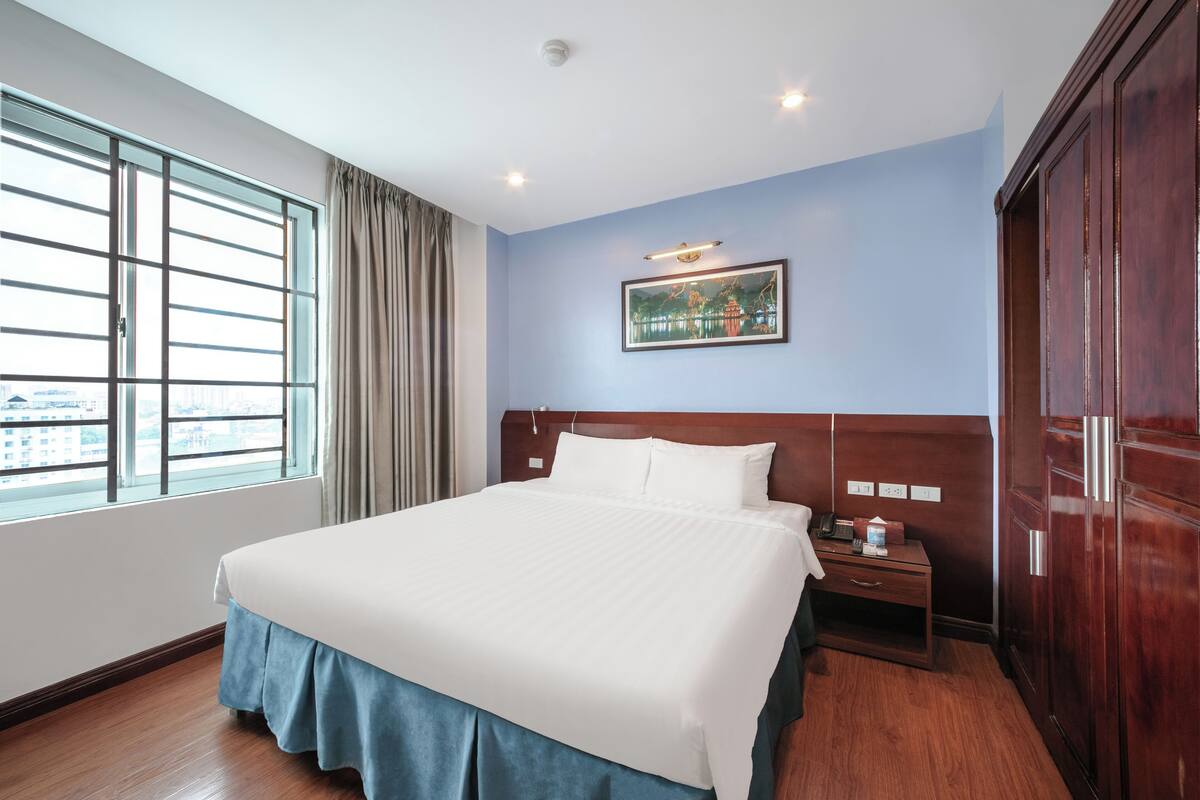 Une chambre d’hôtel avec un grand lit, une table de chevet, une armoire, une fenêtre avec des rideaux et un tableau accroché au mur.