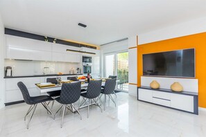 A modern kitchen with white cabinetry, a dining area with black chairs, and a large flat-screen TV mounted on an orange wall.