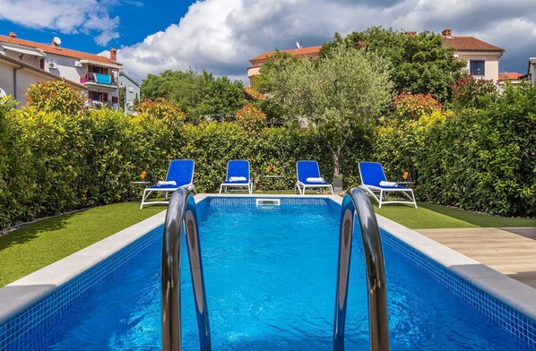 A swimming pool with four blue lounge chairs, surrounded by well-maintained hedges and a clear sky.