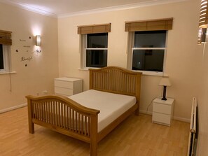 A bedroom with a wooden bed, two bedside tables, a nightstand, a wall-mounted lamp, and a window with a view. - Lovely 3-bed Apartment in London (London)