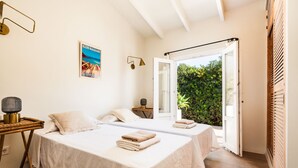 Una habitación con dos camas, una mesita de noche con lámpara, un cartel en la pared y una puerta abierta que da a un jardín.