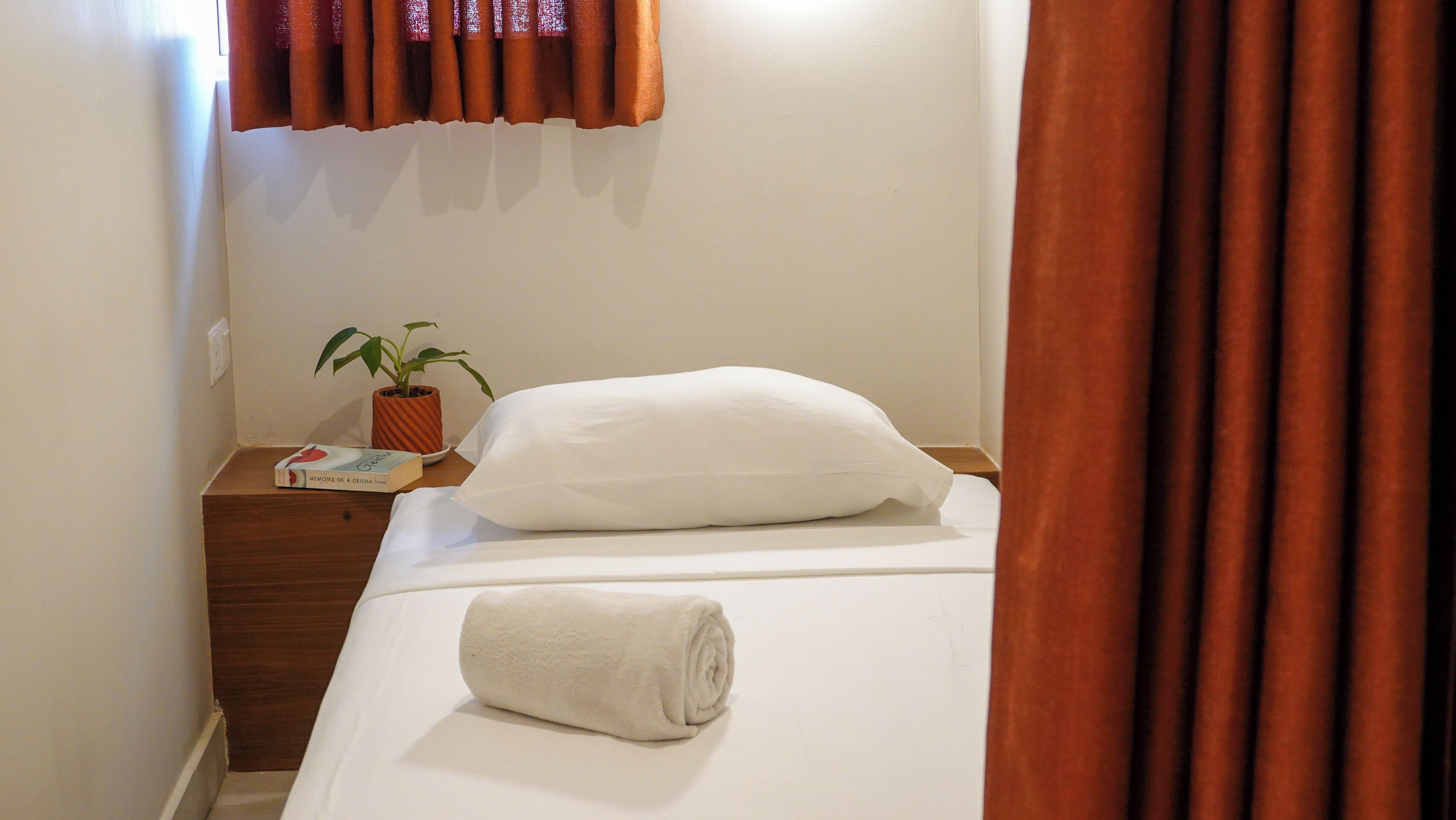Una cama bien hecha con una toalla enrollada, un almohadón blanco, una planta pequeña en maceta y un libro sobre una mesita de noche de madera.
