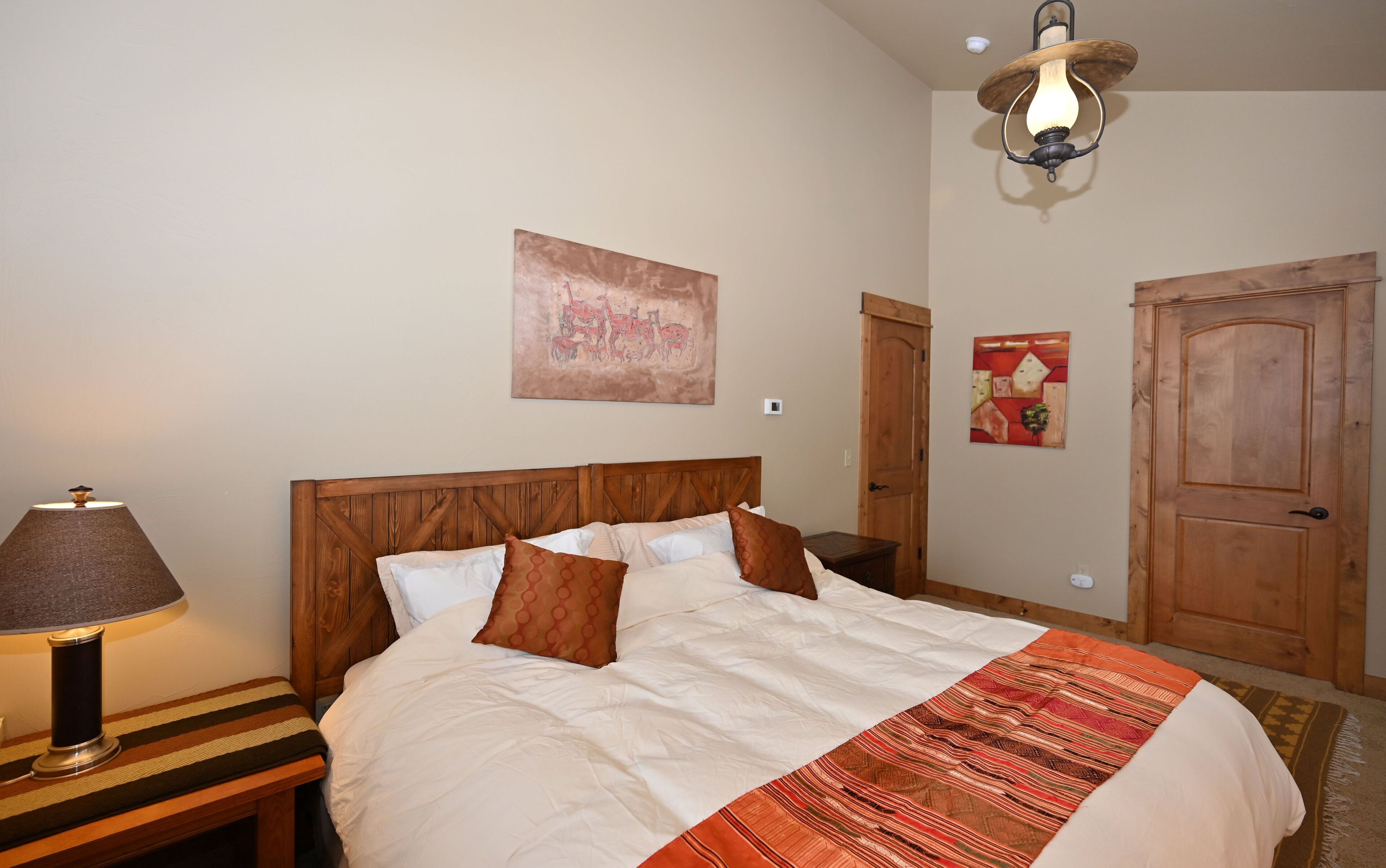 A bedroom with a wooden headboard, a bed with white and brown bedding, a lamp on a bedside table, and a painting on the wall.