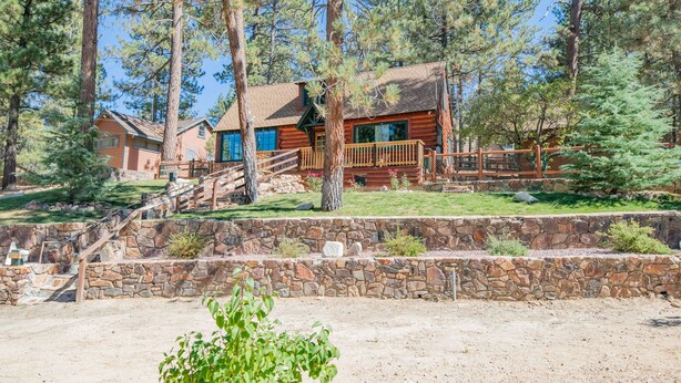 A rustic cabin with a stone retaining wall, wooden deck, and a clear view of the surrounding forest. - Big Bear Retreat - 2 Fireplaces, Deck, Barbecue, Fenced Yard (Big Bear Lake)