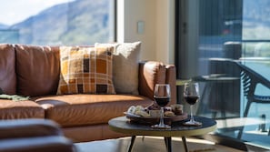 A modern living room with a leather sofa, a round table with a wine glass and snacks, and a view of mountains through a large window. - Woods Villa Tui (Queenstown)