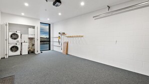 A laundry room with stacked washers and dryers, a window with a view of a lake, and a gray carpeted floor. - Woods Villa Tui (Queenstown)