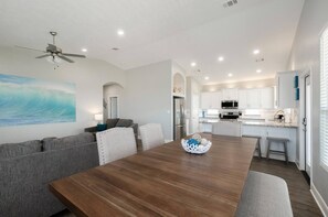 A modern living room with a wooden dining table, grey sofas, and a ceiling fan.