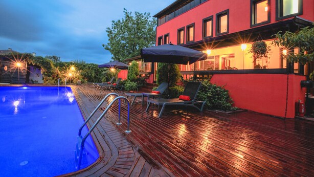 Ein Schwimmbecken mit Liegestühlen und Sonnenschirmen, ein rotes Gebäude mit Fenstern und ein Holzdeck.