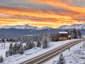 A snow-covered landscape with a road, a two-story building, and snow-capped mountains in the background. - The Hilltop Suite — Mt Massive Views | Pet OK | EV Charger | Sleeps 4 (Leadville)