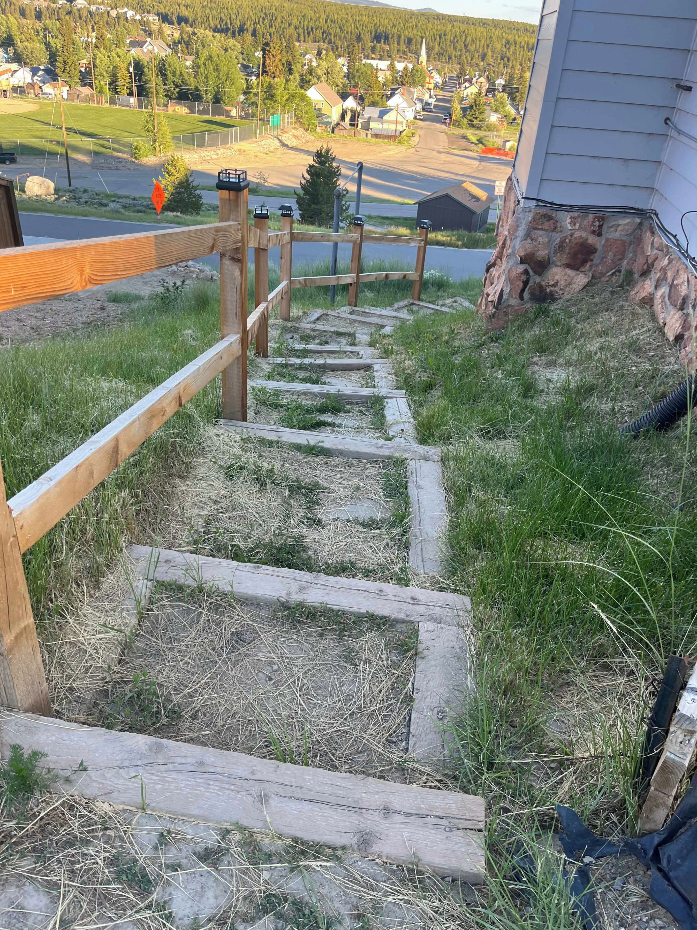 Eine Holztreppe führt zu einem Haus mit Steinunterbau, umgeben von Gras und Blick auf eine Wohngegend.