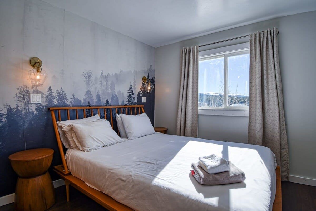 A bedroom with a bed, a nightstand, a wall-mounted lamp, a window with curtains, and a view of the outdoors.