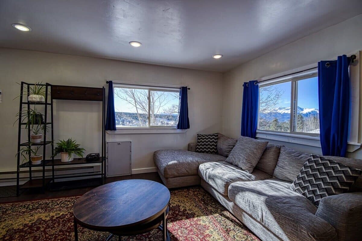 A living room with a sofa, a small table, a plant shelf, and a window with curtains.
