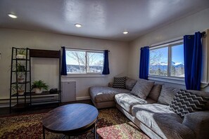 Ein Wohnzimmer mit einer Couch, einem kleinen Tisch, einem Pflanzenregal und einem Fenster mit Vorhängen.