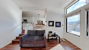 A modern living room with a grey sofa, wooden flooring, a staircase leading to a kitchen, and a large window offering a view of a snowy landscape. - Spacious 2 Bedroom Home with Pool, 3 Hot Tubs, Sauna! by RedAwning (Mammoth Lakes)