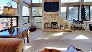 A spacious living room with a fireplace, a television mounted above it, and large windows offering a view of the outdoors. - Deluxe Condo with Fireplace (Unit 623 at 1849) (Mammoth Lakes)