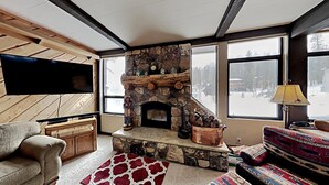 Ein gemütliches Wohnzimmer mit Kamin aus Stein, einem Flachbildfernseher und großen Fenstern mit Blick auf eine verschneite Landschaft.