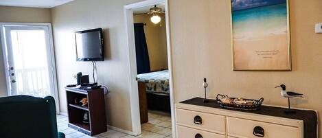 A room with a television, a desk with drawers, a chair, a bed with a curtain, and a framed picture on the wall.
