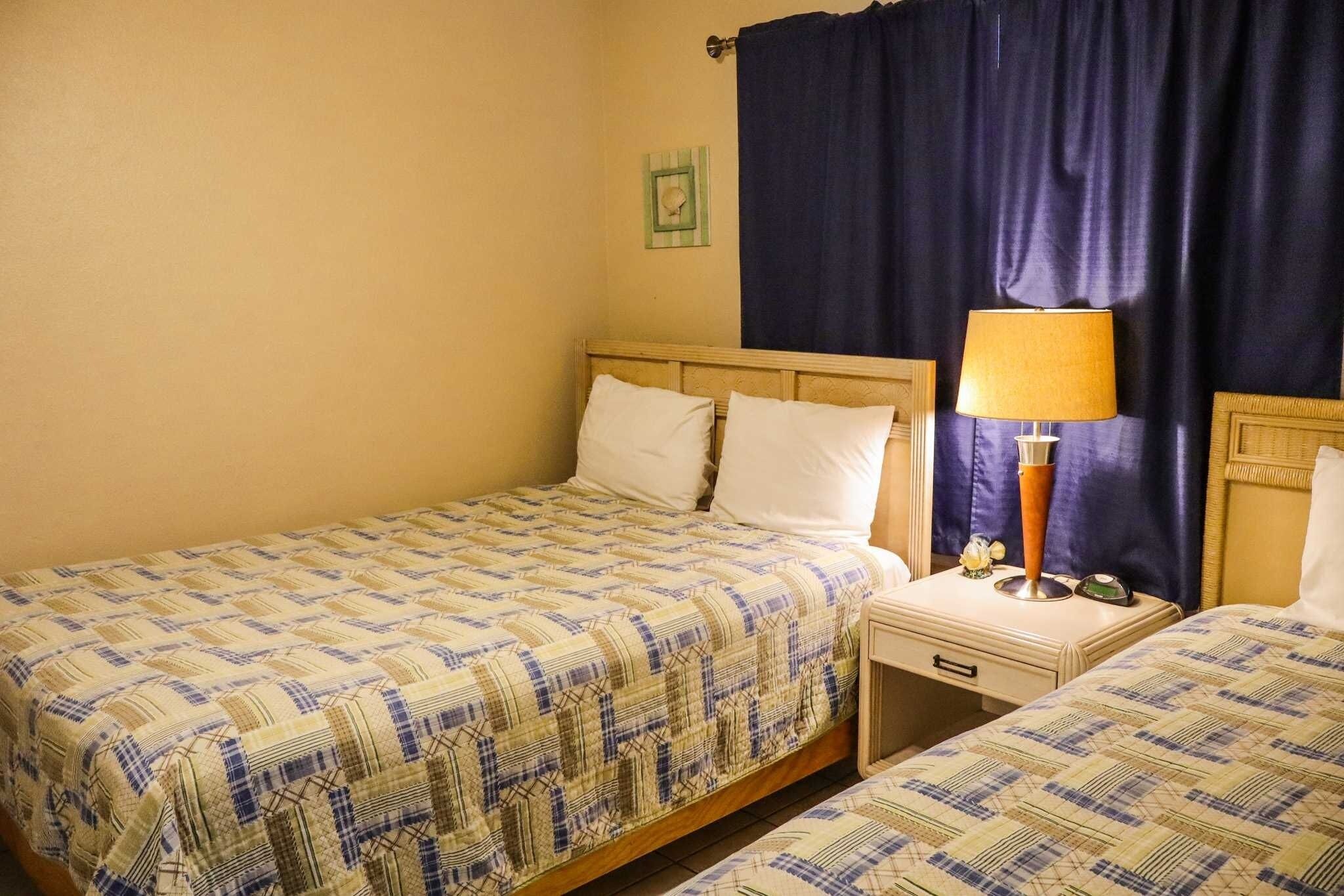 A hotel room with two single beds, a nightstand with a lamp, and a small table with a clock.