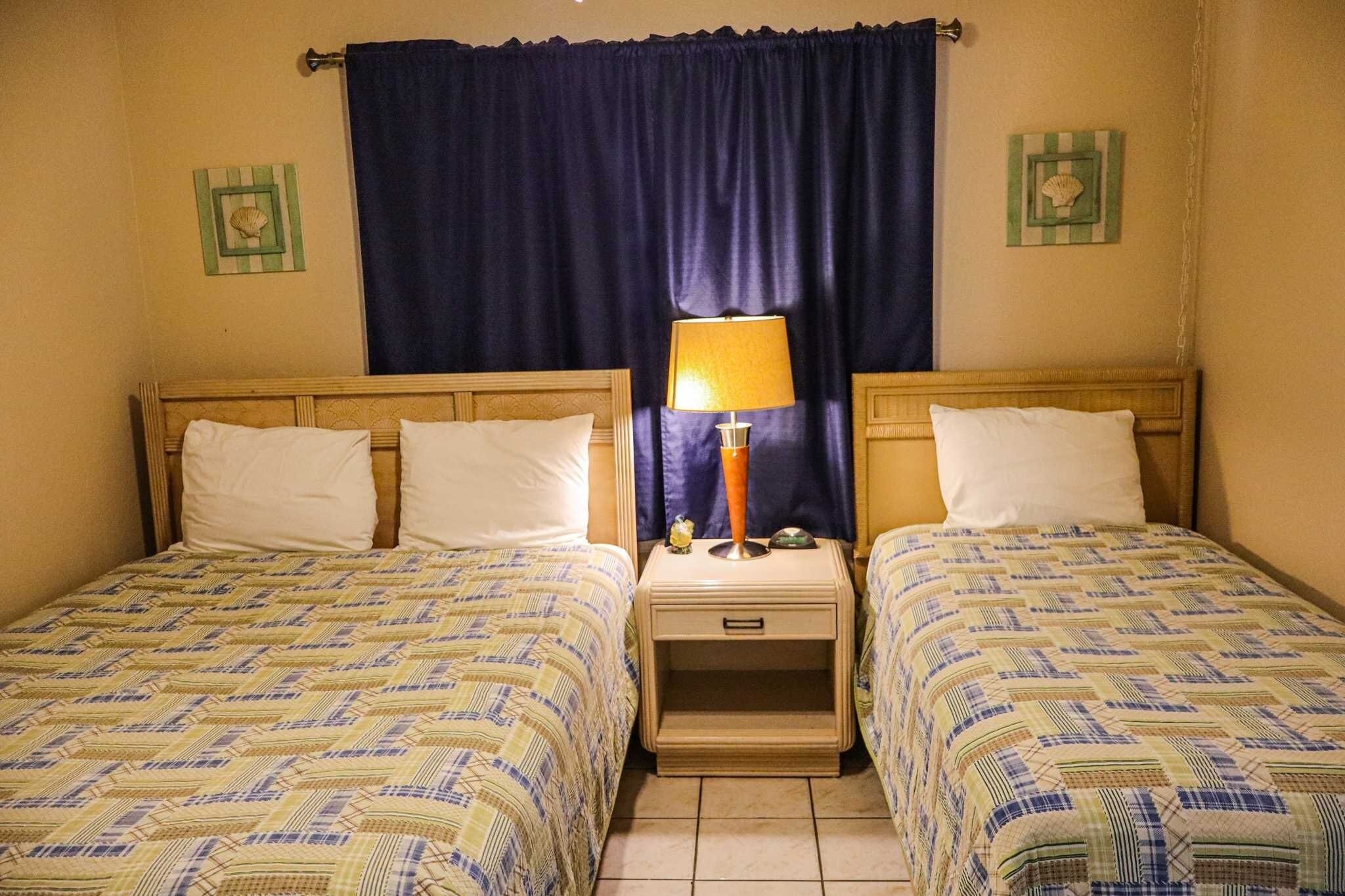 Una habitación con dos camas, cada una con una colcha estampada, una mesita de noche con lámpara y una cortina.