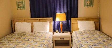 Una habitación con dos camas, cada una con una colcha estampada, una mesita de noche con lámpara y una cortina.