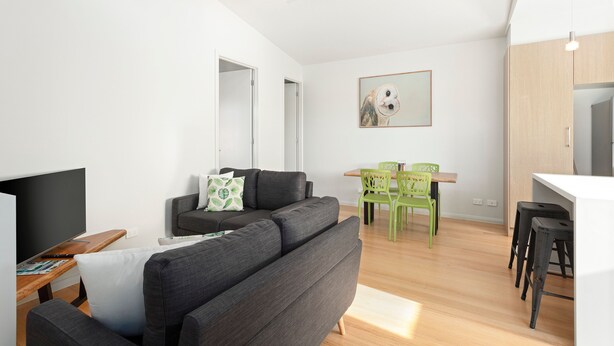 A modern living room with a grey sofa, a flat-screen TV, and a dining area with a wooden table and green chairs. - Bay & Relax (Apollo Bay)