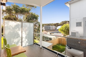 A balcony with a glass door, a wooden table, and chairs, overlooking a garden and buildings.