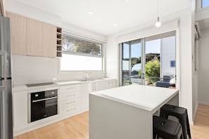 A modern kitchen with a central island, built-in oven, and a large window. - Bay & Relax (Apollo Bay)