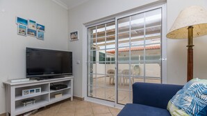 A living room with a flat-screen TV on a white stand, a blue sofa with a patterned pillow, and a sliding glass door leading to an outdoor area with chairs.