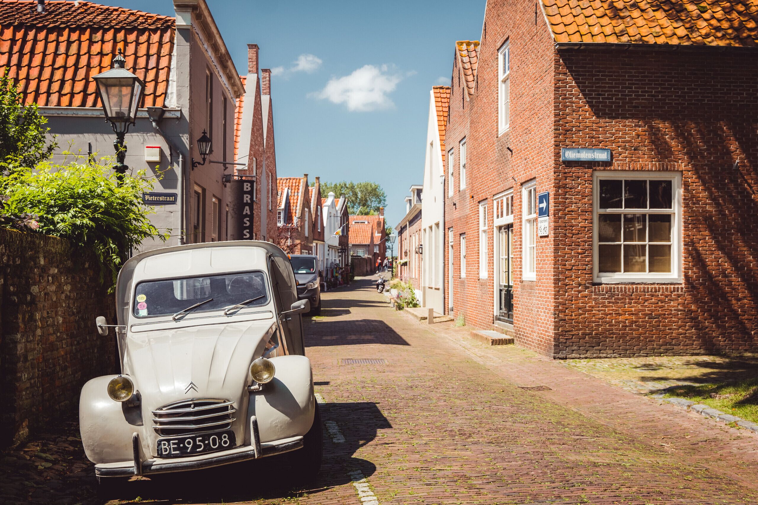 A narrow cobblestone street lined with traditional brick buildings, a vintage car parked on the side, and a clear blue sky.