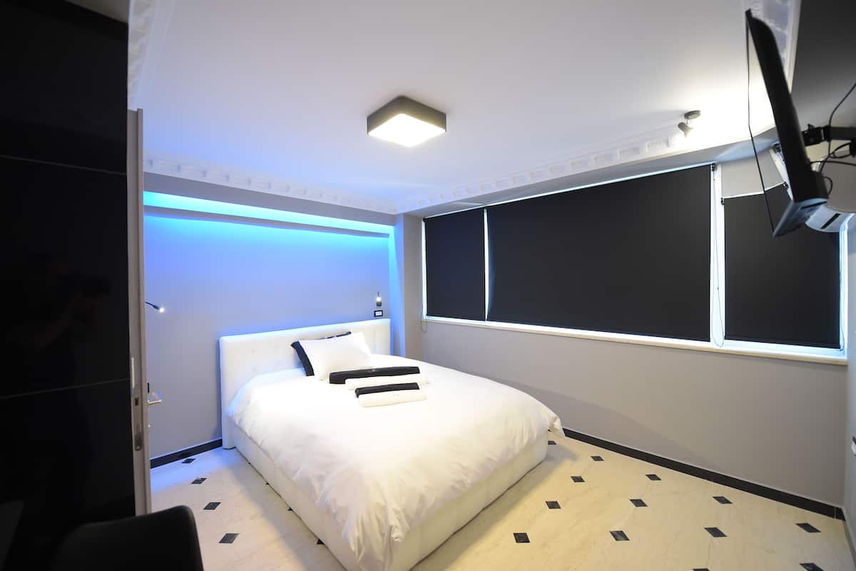 Un dormitorio moderno con una cama grande, un televisor de pantalla plana montado en la pared y un ventanal con cortinas opacas.