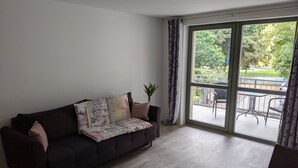 A modern living room with a dark sofa, a patterned throw, and a view of a balcony with outdoor furniture. - Vacations in the historic city center of Zittau - IO19 / 1L (Zittau)