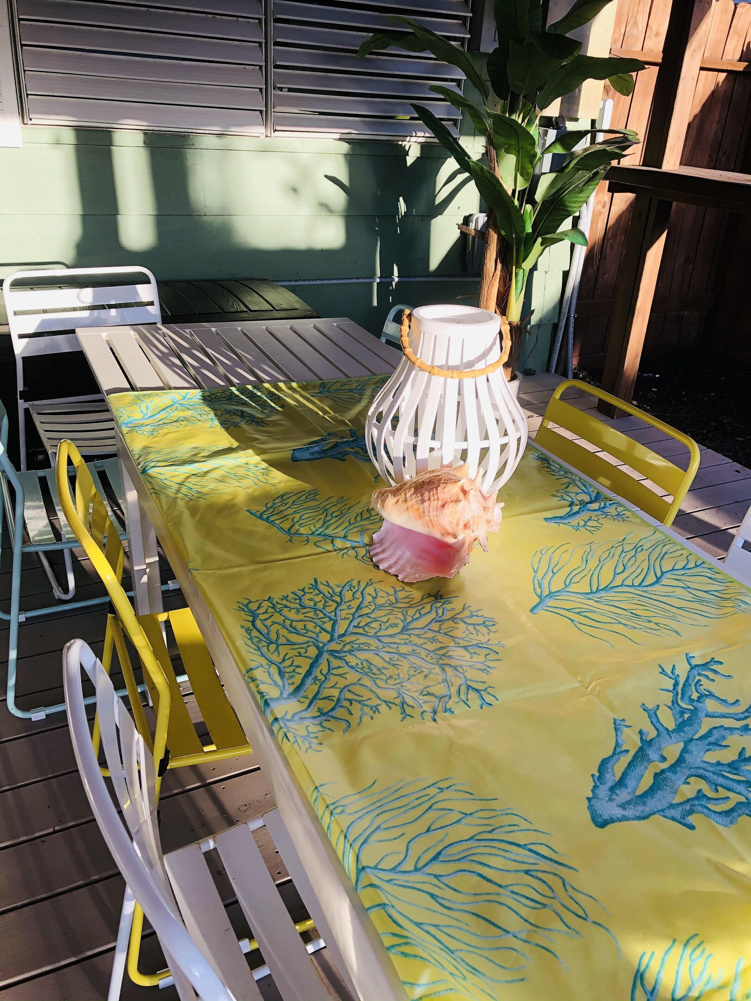 Une table dressée à l’extérieur, recouverte d’une nappe jaune, avec des chaises blanches et une lanterne blanche.