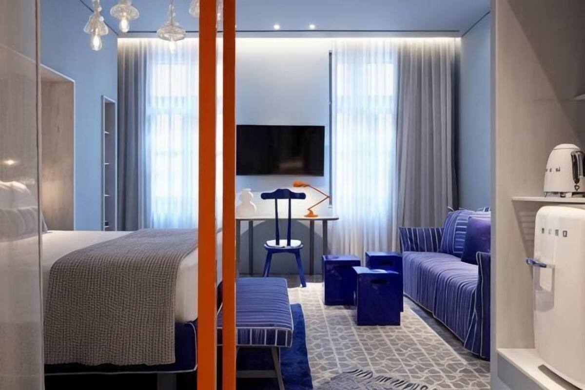 A hotel room with a bed, a desk, a chair, a sofa, a TV, and a minibar.