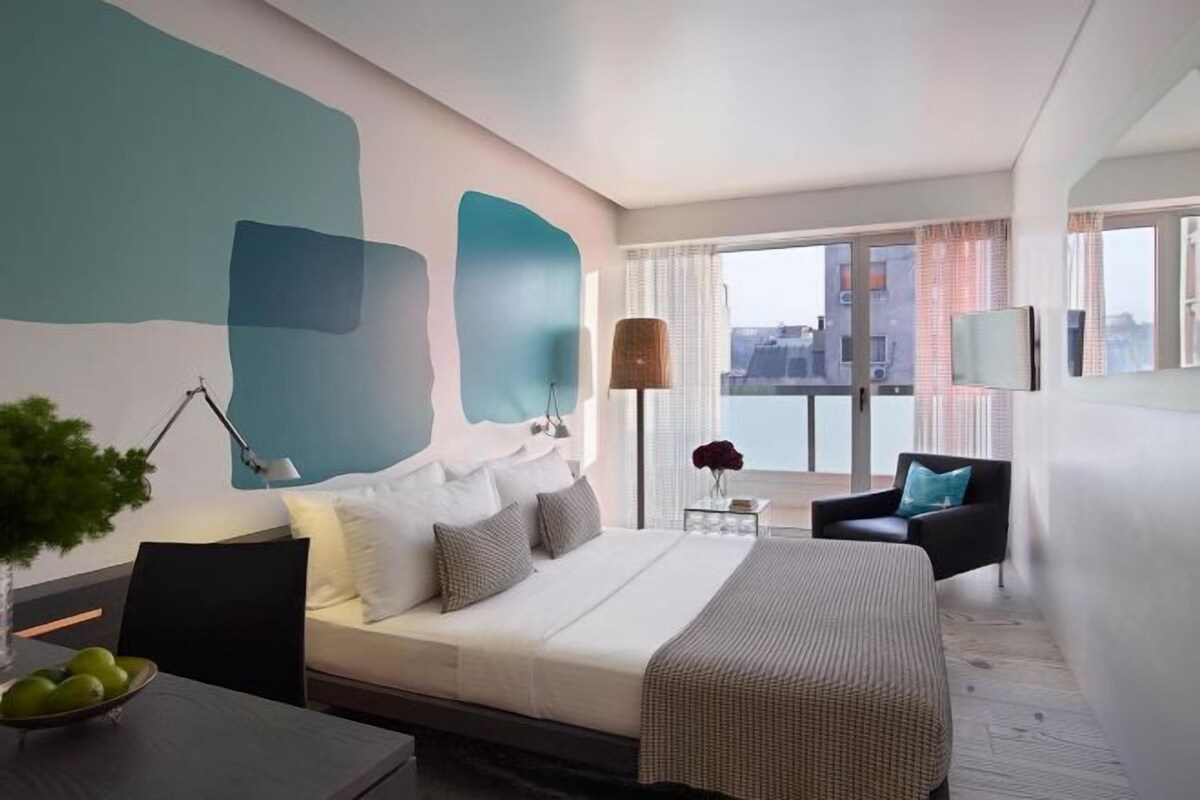 Een moderne hotelkamer met een groot bed, een bureau met lamp, een stoel en uitzicht op de stad door grote ramen.