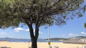 Eine von Bäumen gesäumte Promenade mit einem Sandstrand, blauem Himmel und weiter Ansicht einer Küstenstadt.