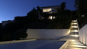 Ein Schwimmbad mit Holzterrasse, umgeben von einer weißen Mauer und einer Treppe, die zu einem Haus führt.